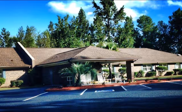 Exterior view of a single-story brick building with a covered entrance featuring rocking chairs. The building is surrounded by trees and shrubs, with a parking area in front including a handicapped parking space.