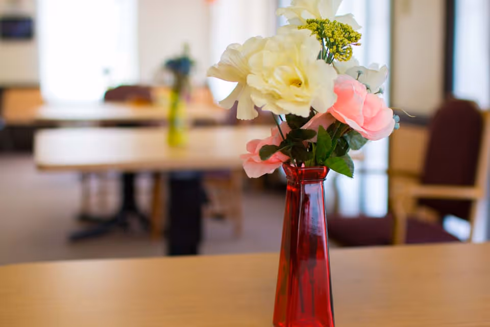 A red vase holding white and pink flowers sits on a table in a blurred communal dining room.