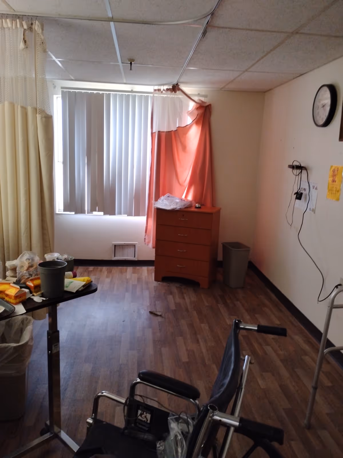 A sparse rehabilitation room with a wheelchair, dresser, table, curtains, and medical supplies.