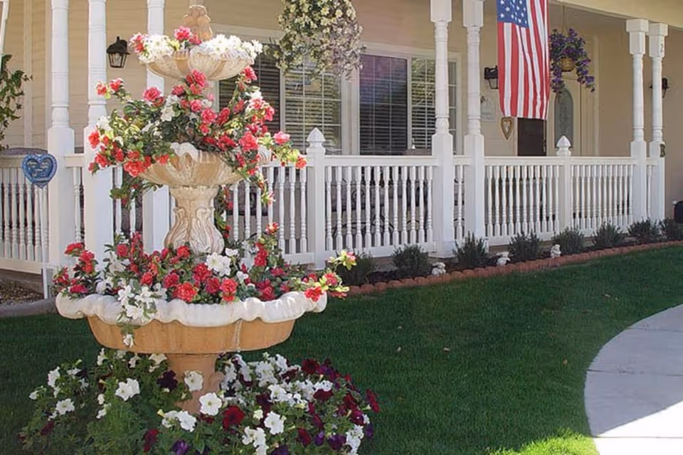 Tiered stone fountain filled with pink and white flowers on a green lawn in front of a porch with white railing and an American flag.