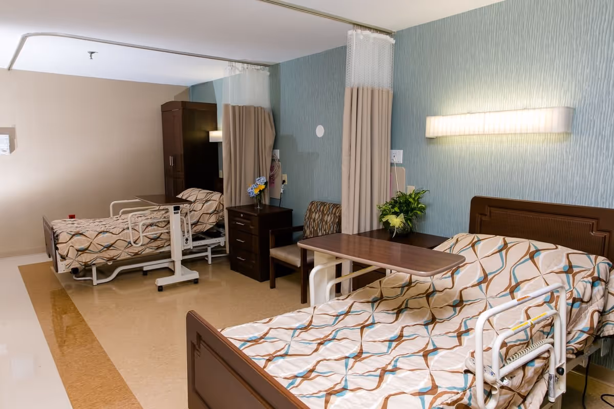 A shared hospital-style room with two beds separated by a privacy curtain. Each bed has a patterned bedspread with brown, blue, and beige colors. There are overbed tables, a wooden nightstand with drawers, a chair, and a potted plant on the nightstand. The walls have a textured blue wallpaper and a mounted light fixture above each bed.