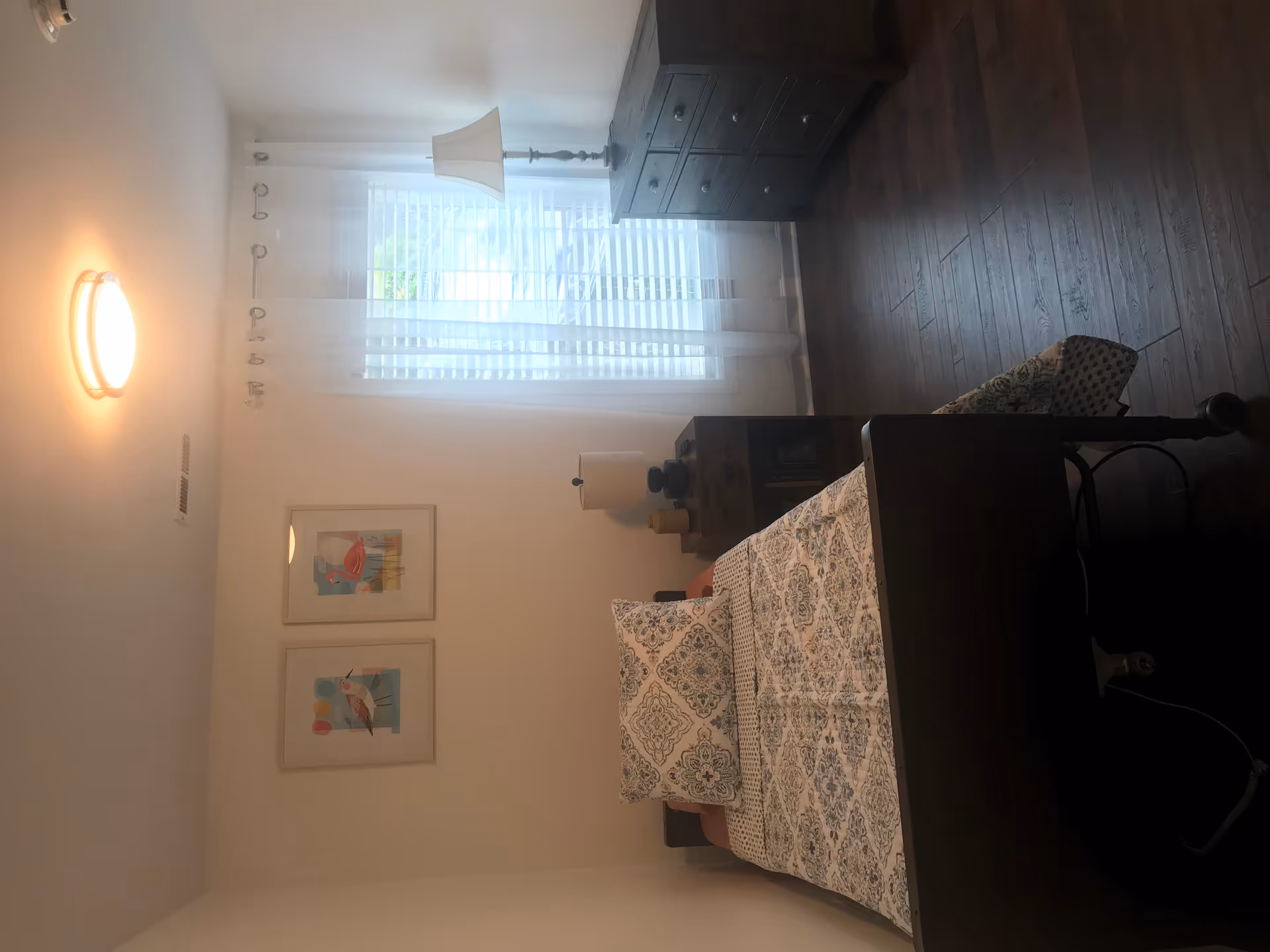 A furnished bedroom in a senior living facility with bed, dresser, nightstand, lamp and window blinds.