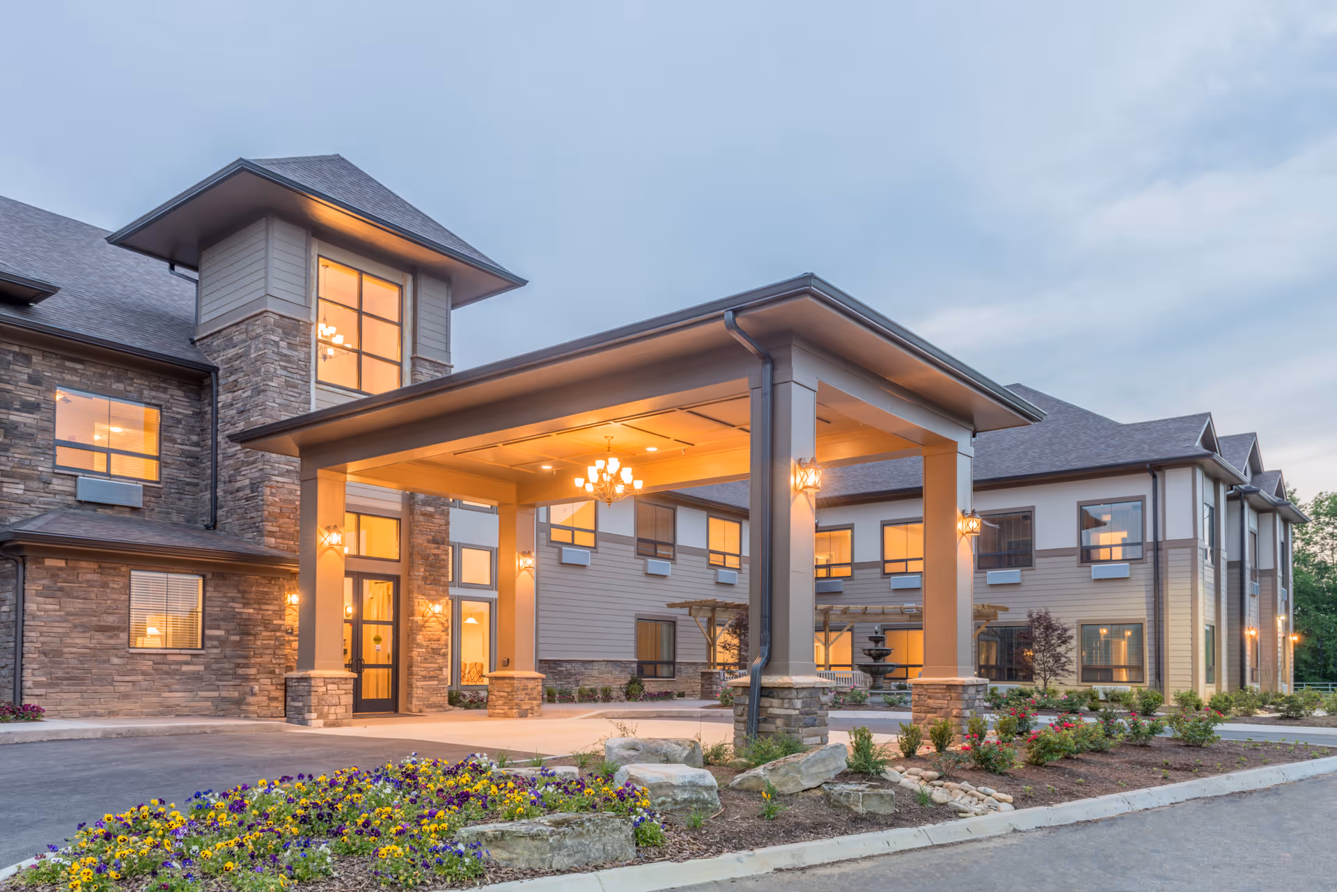 Front entrance of a modern senior living facility with a covered porte-cochere, illuminated windows, and landscaped flower beds.