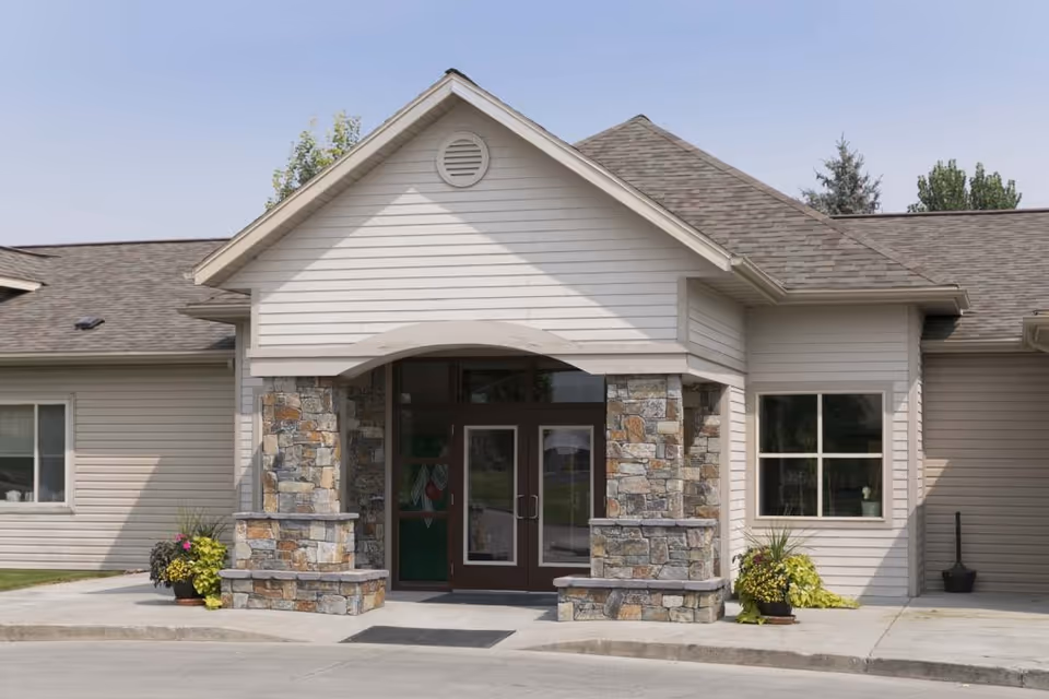 Front exterior view of a single-story building with beige siding and a stone entrance. The entrance has double glass doors with stone pillars on either side and potted plants near the entrance. The roof is shingled and there are windows on either side of the entrance.