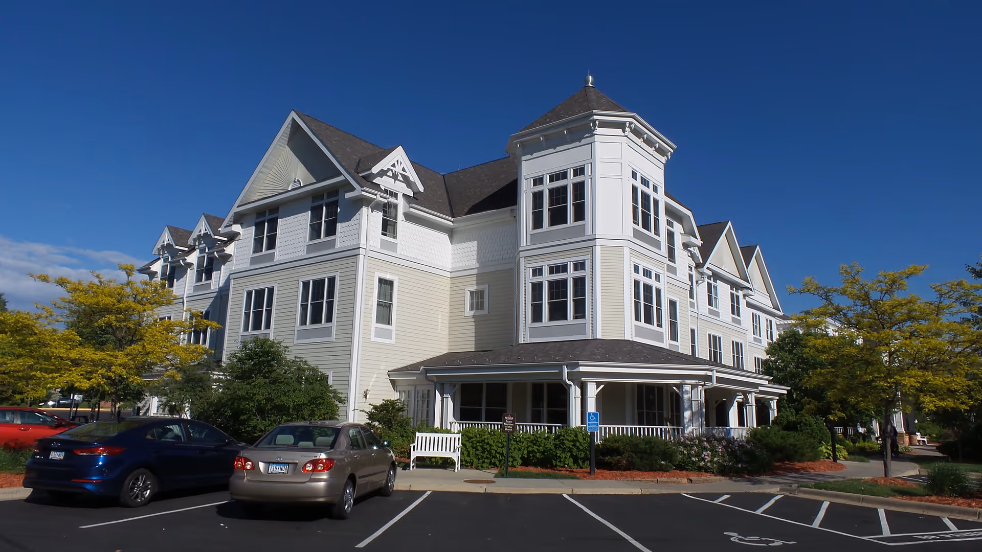 White multi-story senior living building with a front porch, landscaping, and parked cars.