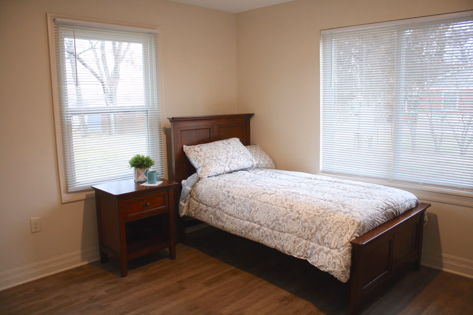 A neat bedroom with a wooden bed, nightstand, and large windows with blinds.