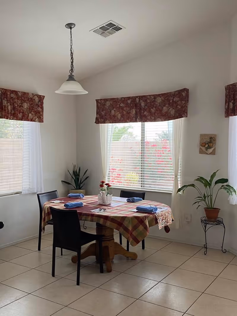 Small dining room with a round table covered by a plaid tablecloth, four chairs, folded blue napkins, potted plants and windows with floral valances.