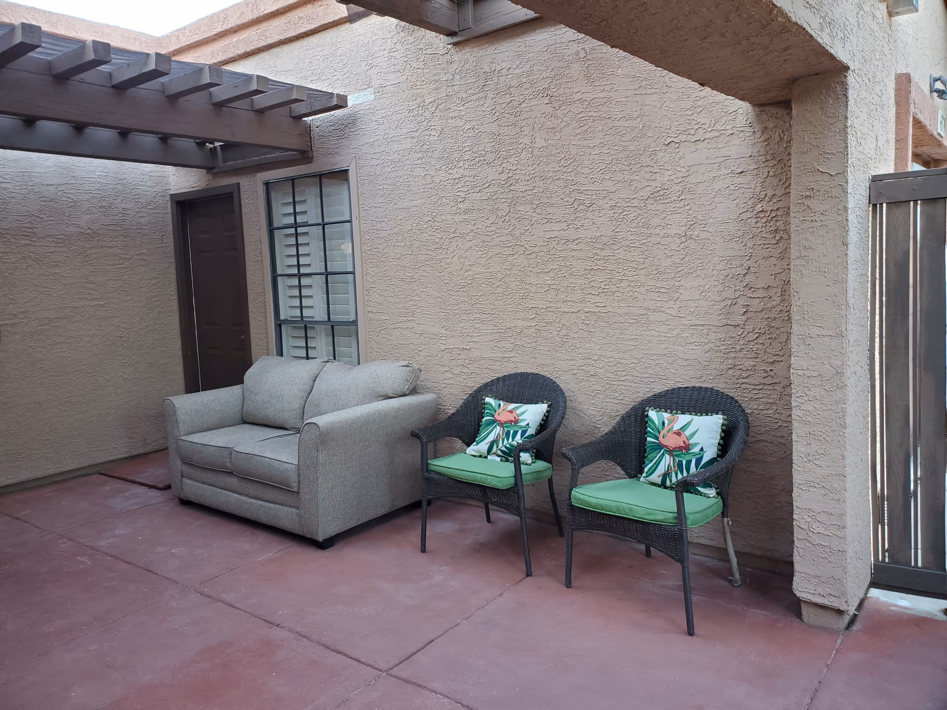 Outdoor patio area with a beige loveseat and two black wicker chairs with green cushions and flamingo-themed pillows, against a textured beige wall with a window and door under a wooden pergola.