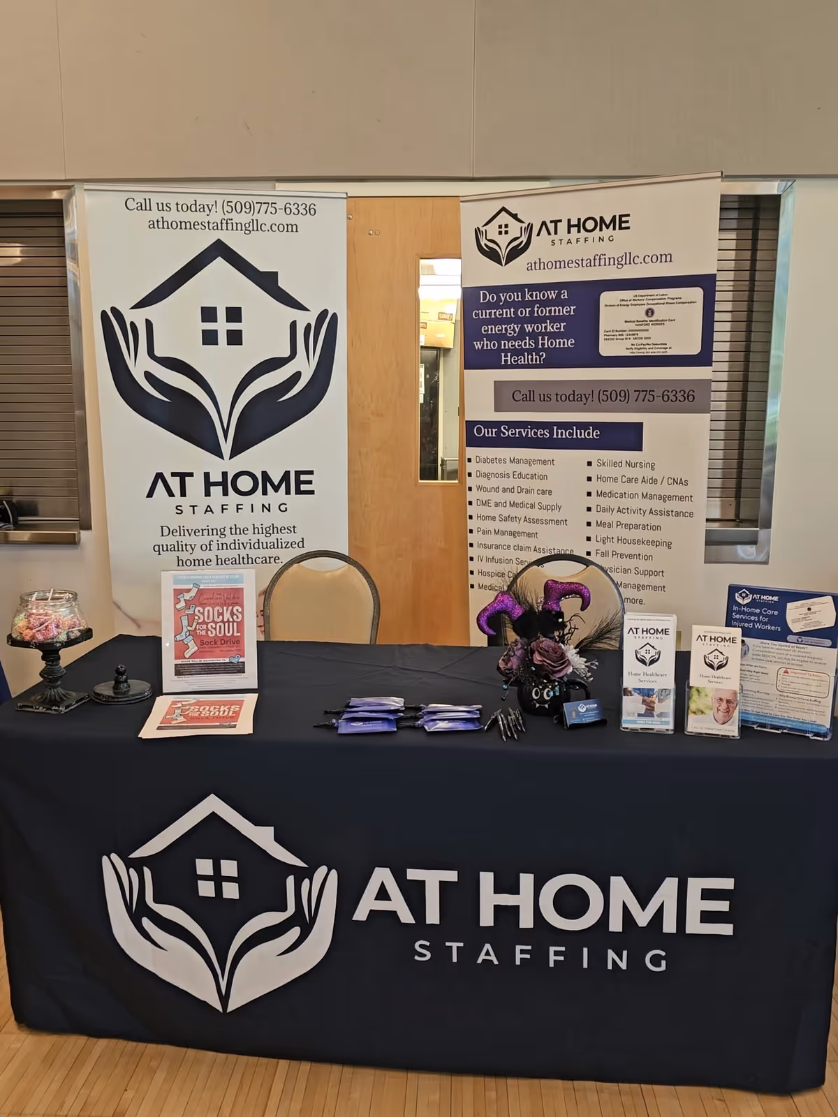 Promotional booth for At Home Staffing with banners, a table, and brochures indoors.