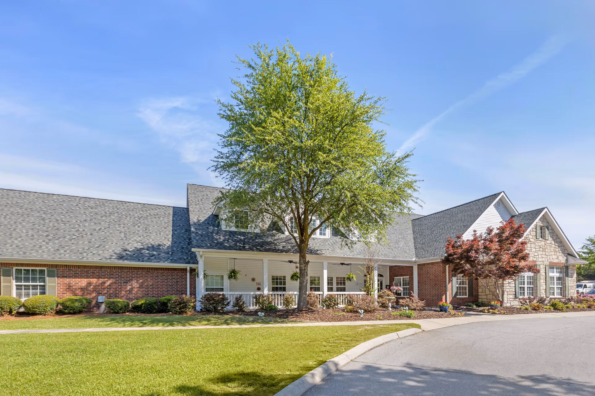 Exterior view of a senior living facility building with a mix of brick and stone facade, a large tree in front, well-maintained landscaping, and a clear blue sky.