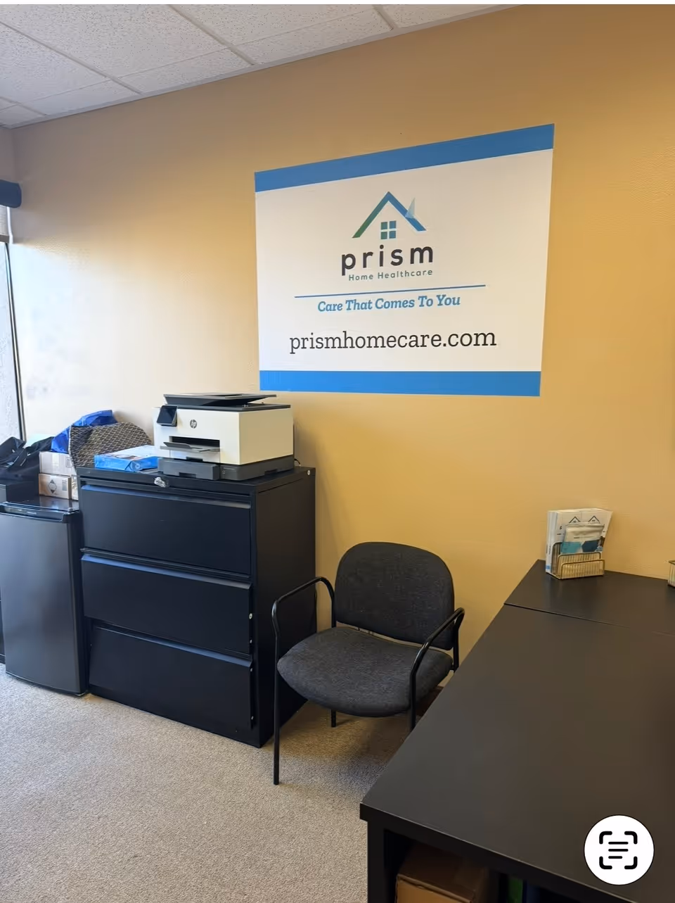 Office corner with printer, filing cabinet, and Prism Home Care sign on the wall.