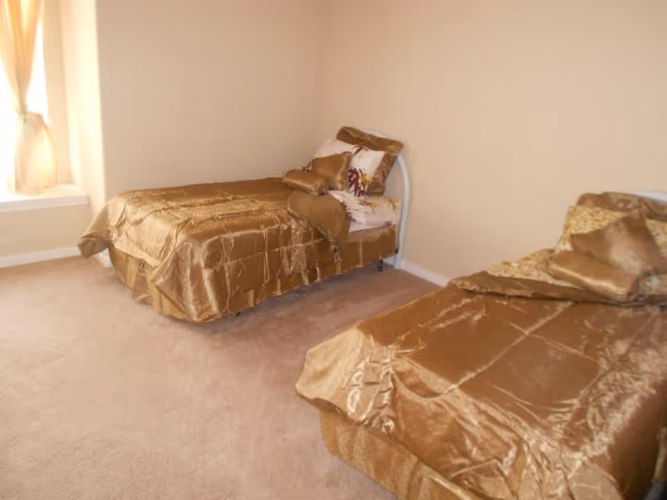 Two twin beds with gold bedspreads in a sparsely furnished bedroom with beige walls and a curtained window.