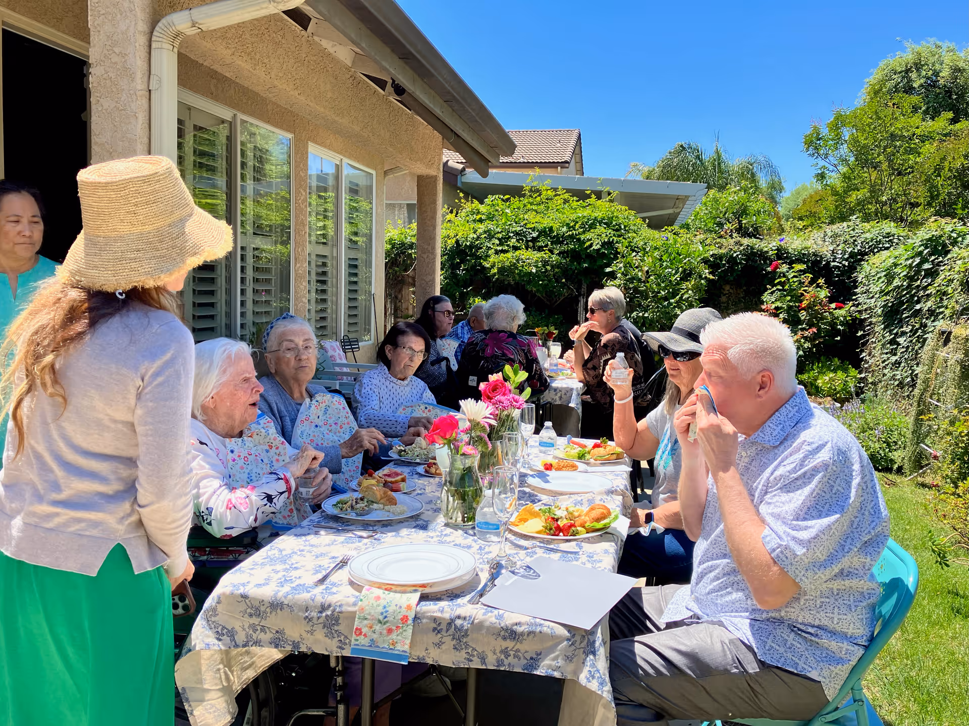 A group of elderly people and caregivers sitting around a long table outdoors, enjoying a meal together in a garden setting with greenery and flowers under a clear blue sky.