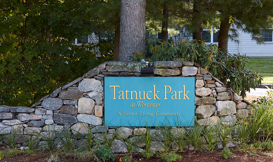 Stone wall sign with a blue plaque that reads 'Tatnuck Park at Worcester A Senior Living Community' surrounded by green plants and trees, with part of a white building visible in the background.