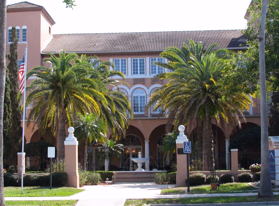 Peach-colored building front with arches, palm trees, a fountain, and an American flag.