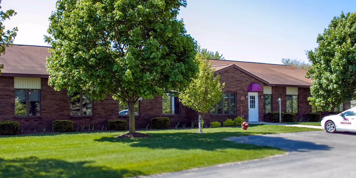 Brick one-story building with trees, a driveway, and a white door with a purple awning.