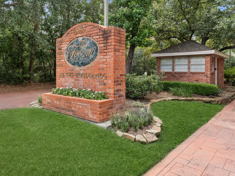 Brick entrance sign reading 'The Forum at The Woodlands' next to a small brick guardhouse surrounded by trees and landscaping.
