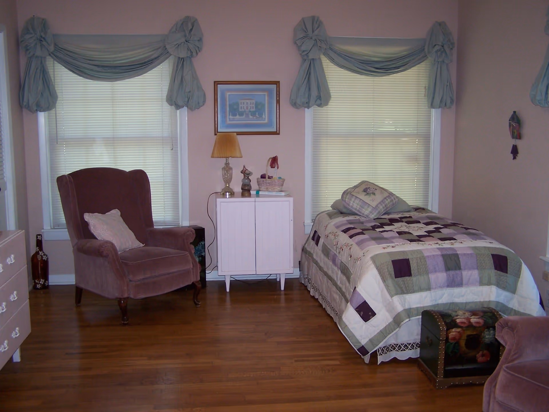 A cozy bedroom with two windows covered by light blue draped curtains. There is a single bed with a patchwork quilt and pillows on the right side, a white cabinet with a lamp and decorative items in the center, and a plush armchair with a cushion on the left side. The room has wooden flooring and light-colored walls.