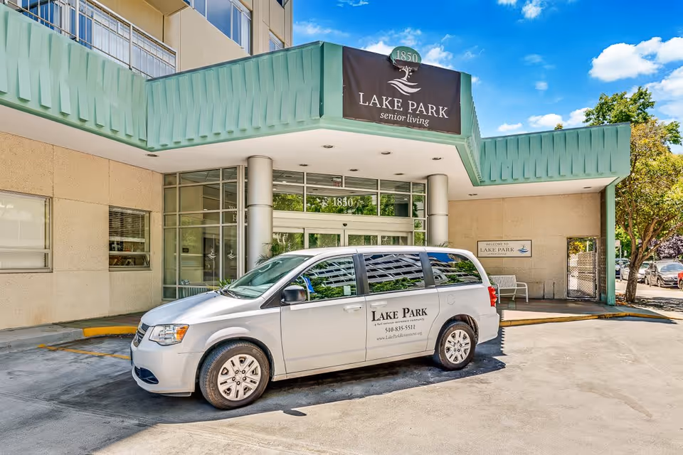 Exterior view of the entrance to Lake Park Senior Living facility with a silver van parked in front. The building has a green awning with the Lake Park Senior Living sign above the entrance. Trees and a clear blue sky are visible in the background.