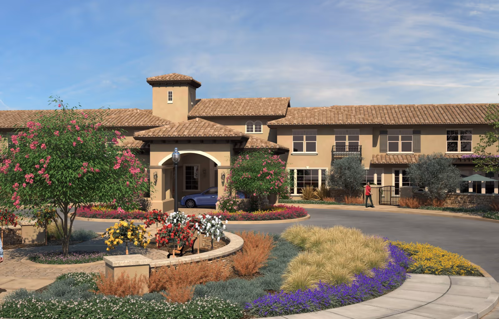Front entrance of a Mediterranean-style senior living building with a circular driveway, landscaped flower beds, and a person walking by.