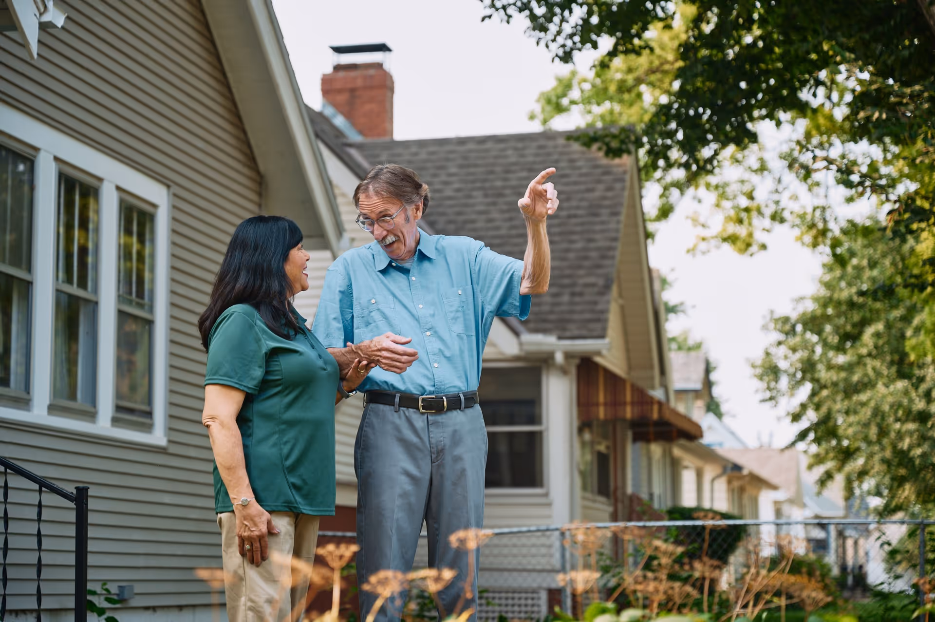 Two seniors stand outside a row of houses, chatting and smiling in a sunny residential area.