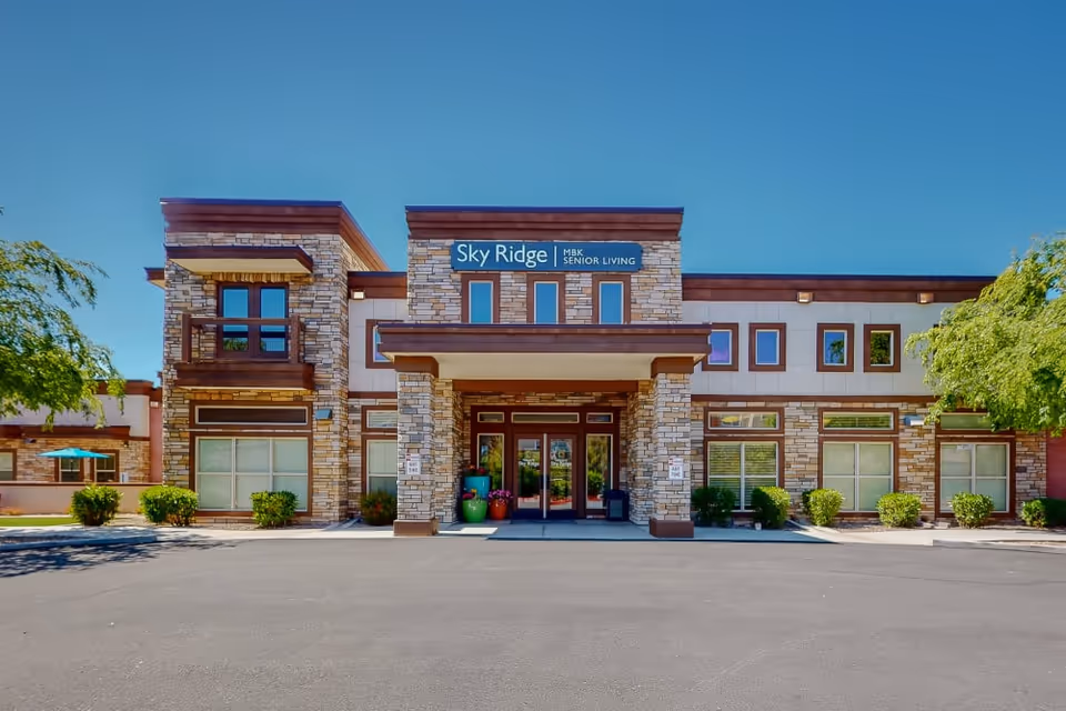 Front exterior view of the Sky Ridge MBK Senior Living facility, featuring a modern two-story building with stone and wood accents, large windows, and a covered entrance with potted plants. The sky is clear and blue, and there are trees and shrubs around the building.