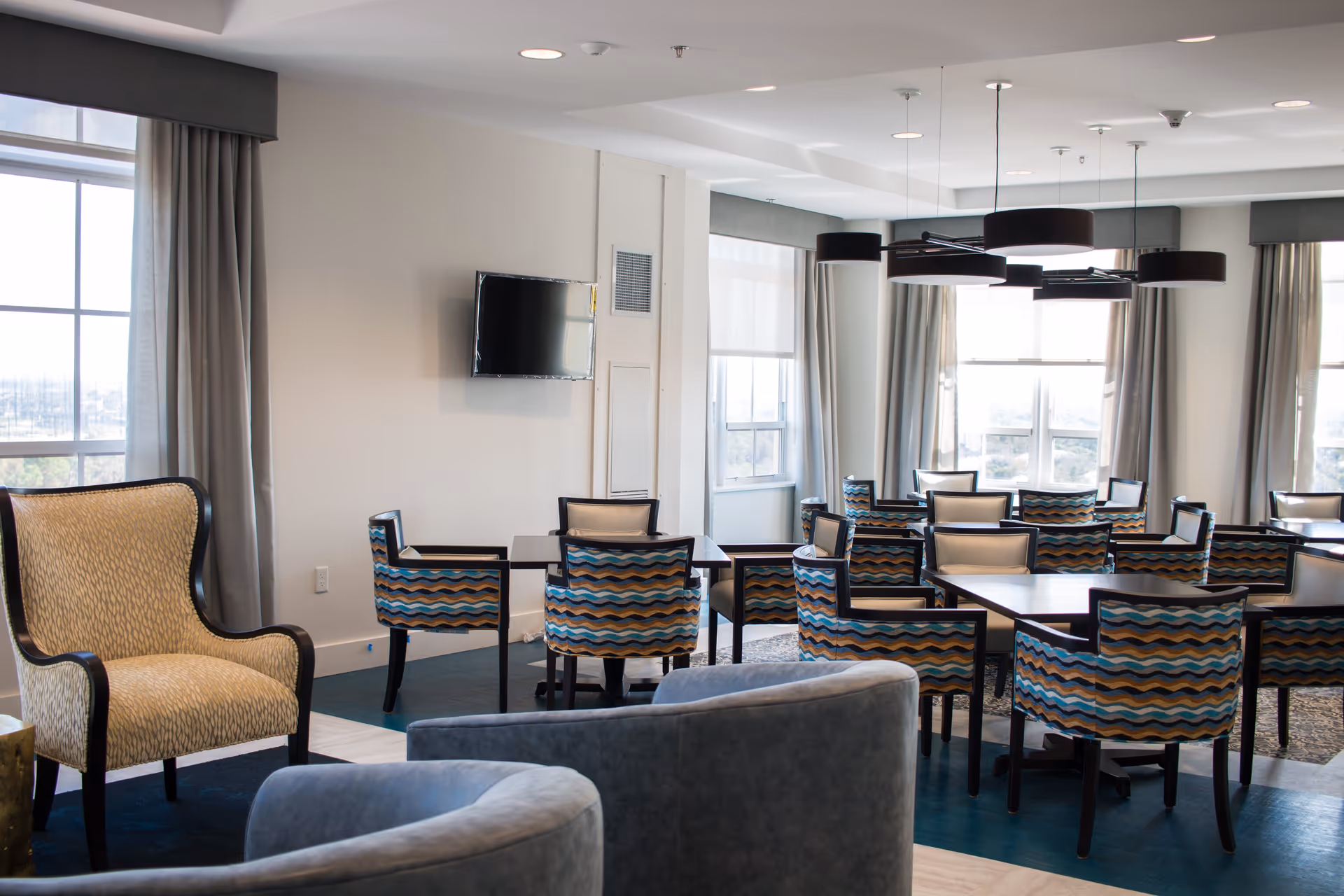Bright communal dining room with patterned chairs, multiple tables, armchairs, large windows, and a wall-mounted TV.