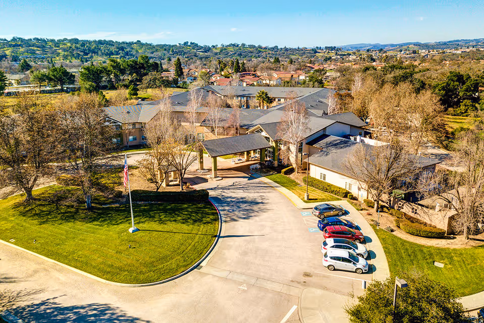 Aerial view of Creston Village Assisted Living and Memory Care facility showing a large building with a covered entrance, surrounded by trees and greenery. Several cars are parked in the parking lot near the entrance, and an American flag is visible on a flagpole on a grassy area in front of the building.