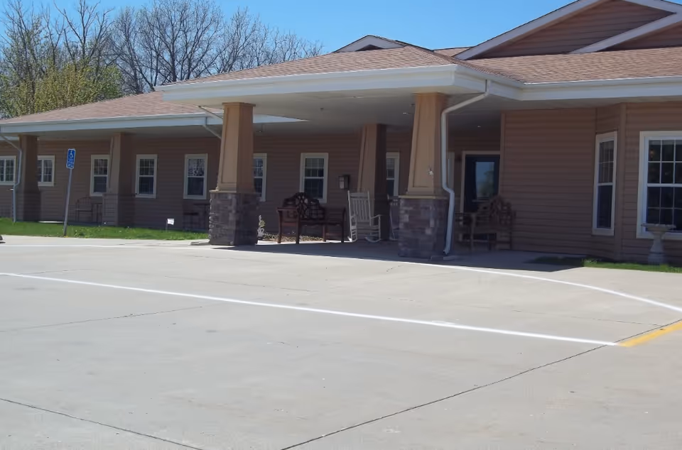Entrance of a single-story senior living facility with a covered drive-up portico, columns, benches and a parking area.