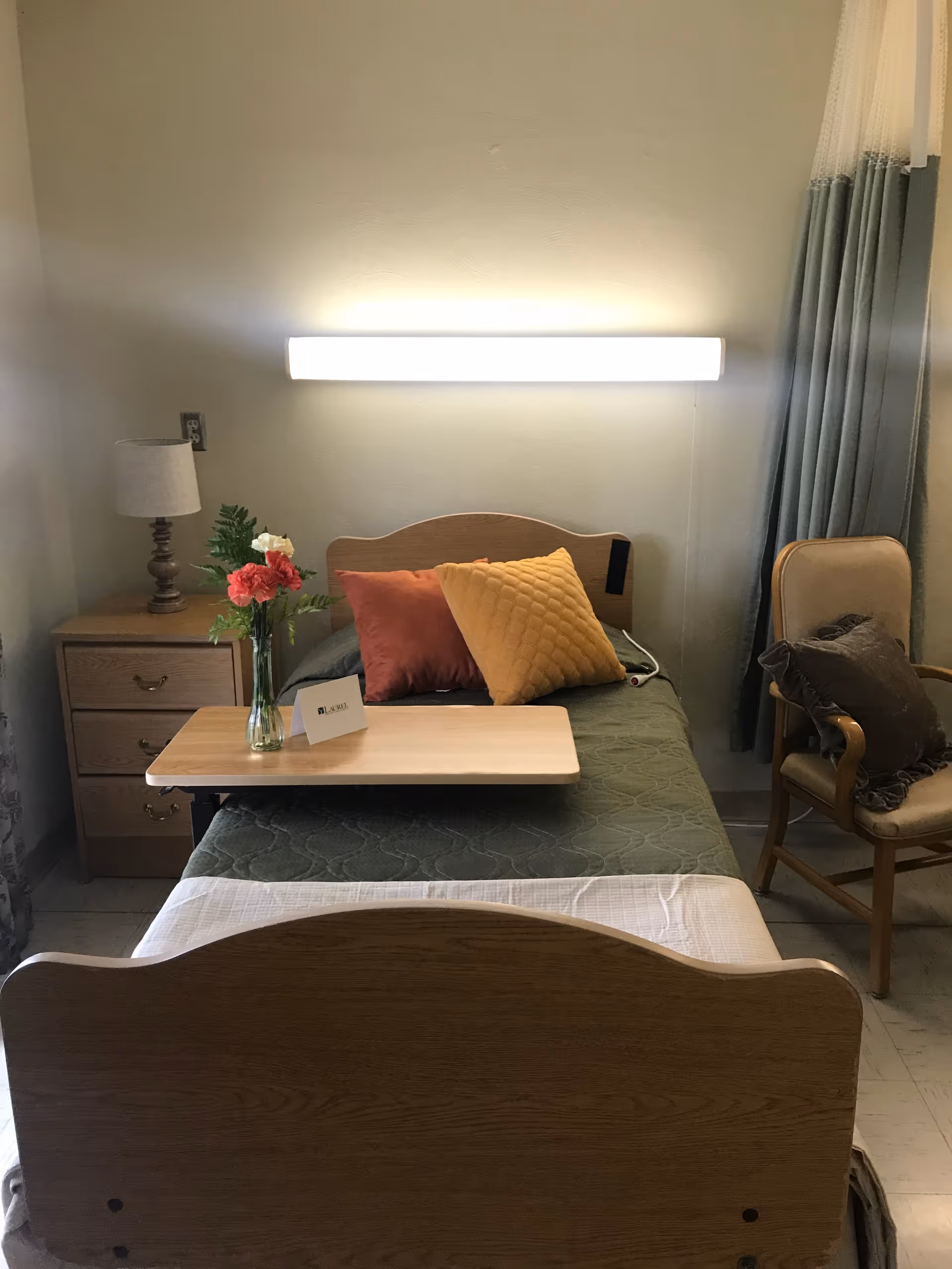 A single bed with a green quilt and two pillows, one orange and one yellow, is positioned against a wall with a bright horizontal light fixture above it. A wooden bedside table with a lamp and a vase of flowers is on the left side of the bed. A wooden chair with a cushion is on the right side near a window with blue and white curtains. A wooden overbed table is placed on the bed with a small card on it.