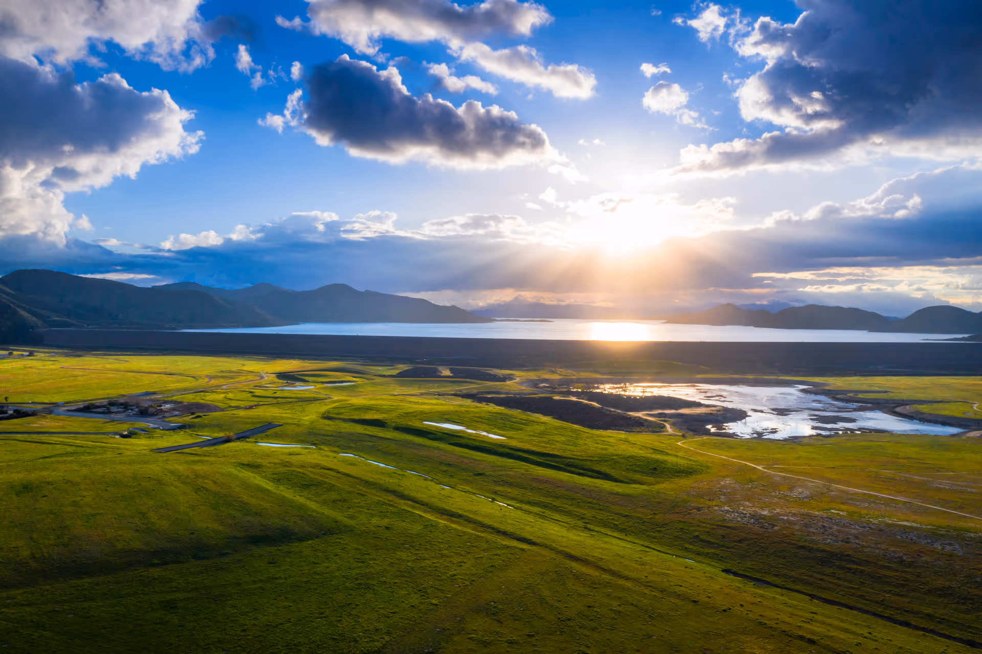 Sunlit valley with green fields, distant mountains, and a reflective body of water.