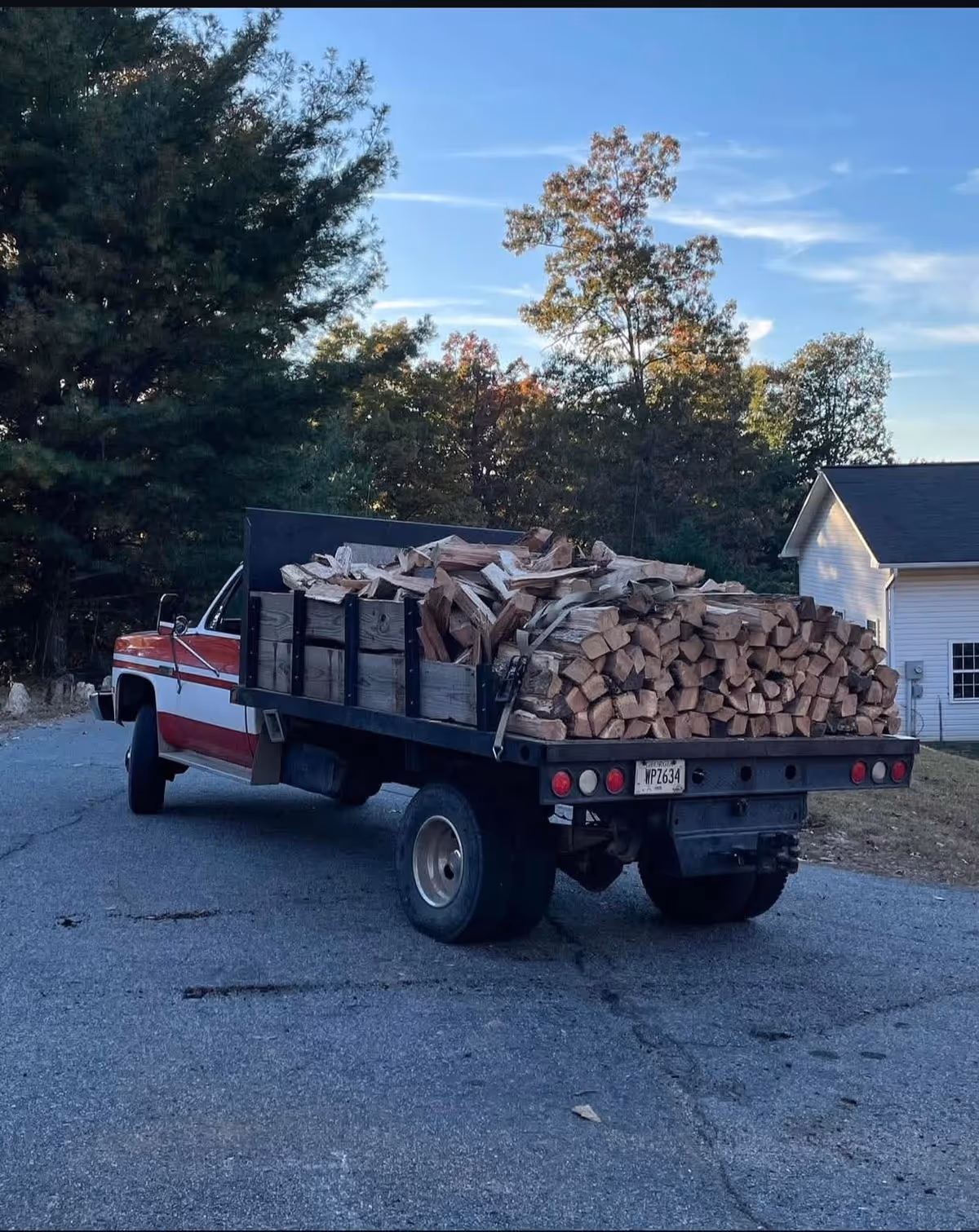 Red-and-white pickup truck with a trailer filled with firewood parked beside trees and a white house.