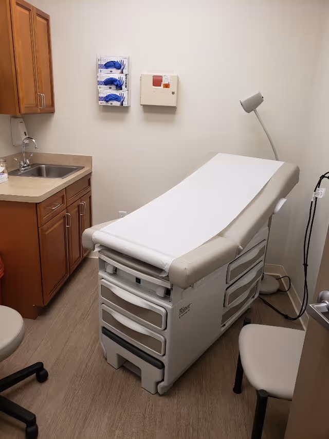 A clinic examination room with a medical exam table, sink, and wooden cabinets.