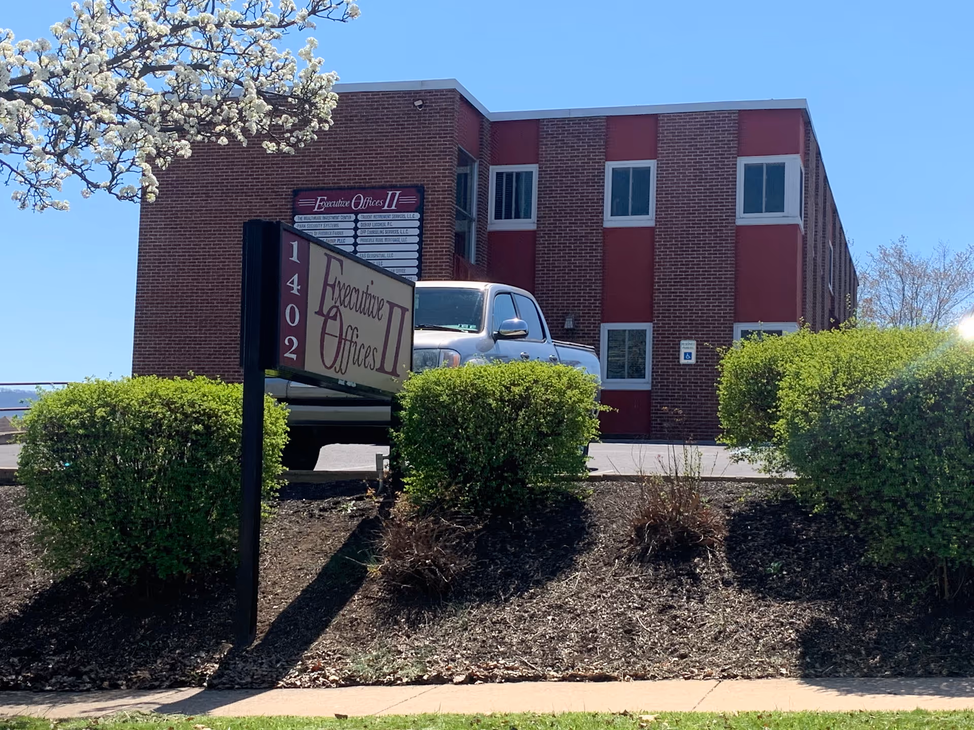 Brick office building facade with sign Executive Offices II, a parked car, and trimmed shrubs.