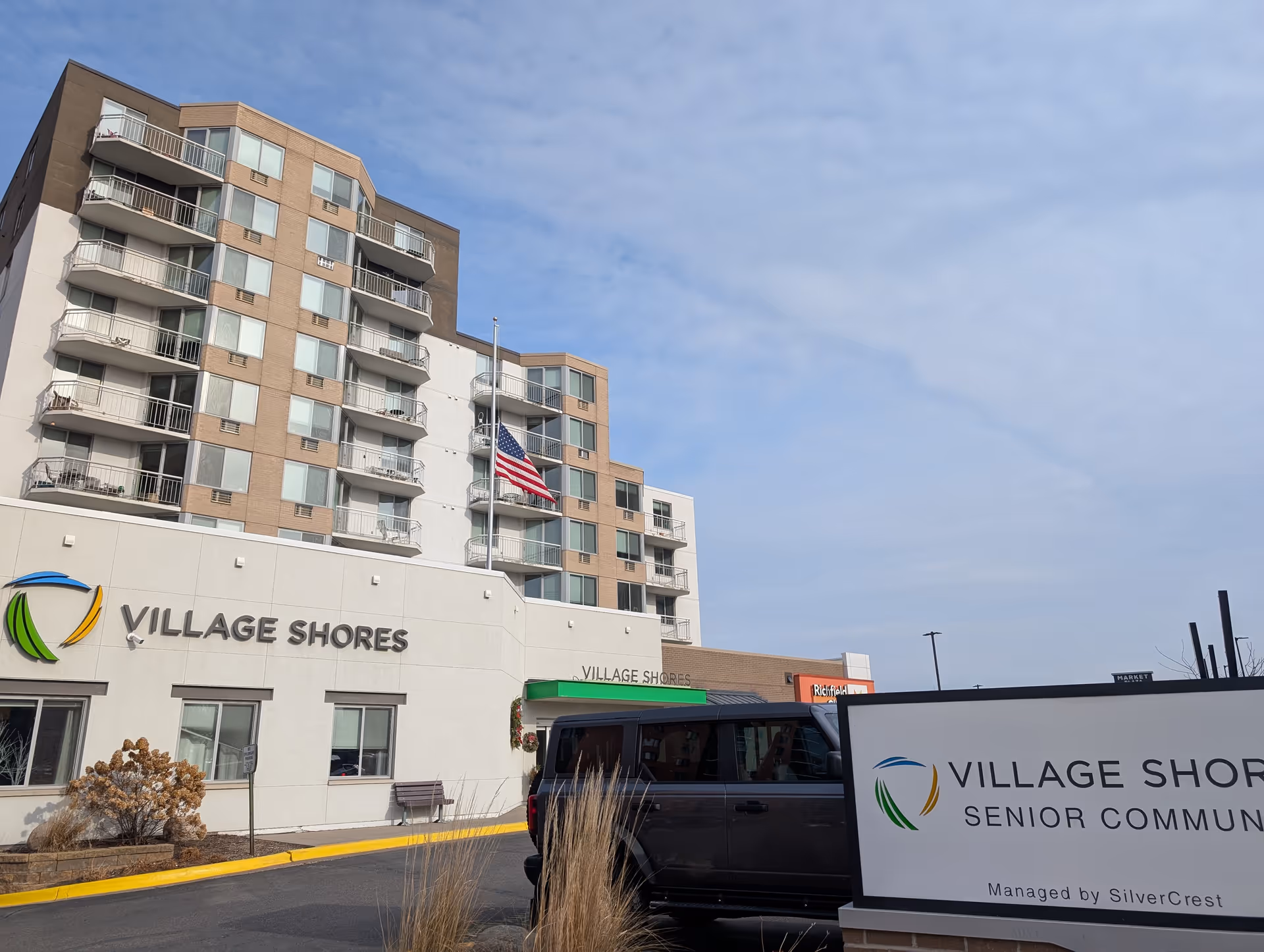 Front exterior of the Village Shores senior living building showing the entrance, signage, balconies, and an American flag.