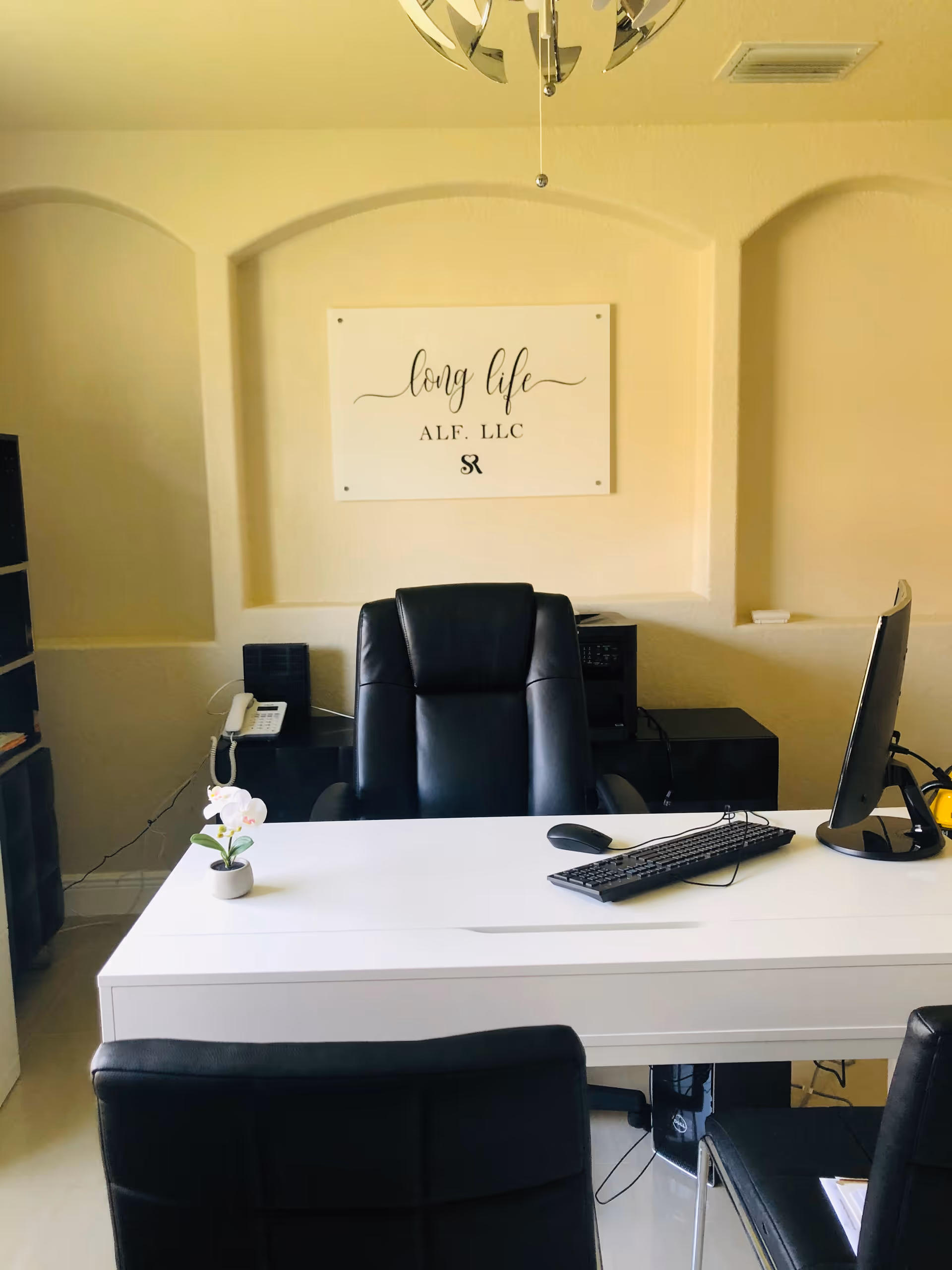 An office space with a white desk, a black leather office chair behind the desk, and two black chairs in front. On the desk are a computer monitor, keyboard, mouse, and a small potted plant. On the wall behind the desk is a sign that reads 'long life ALF, LLC'.