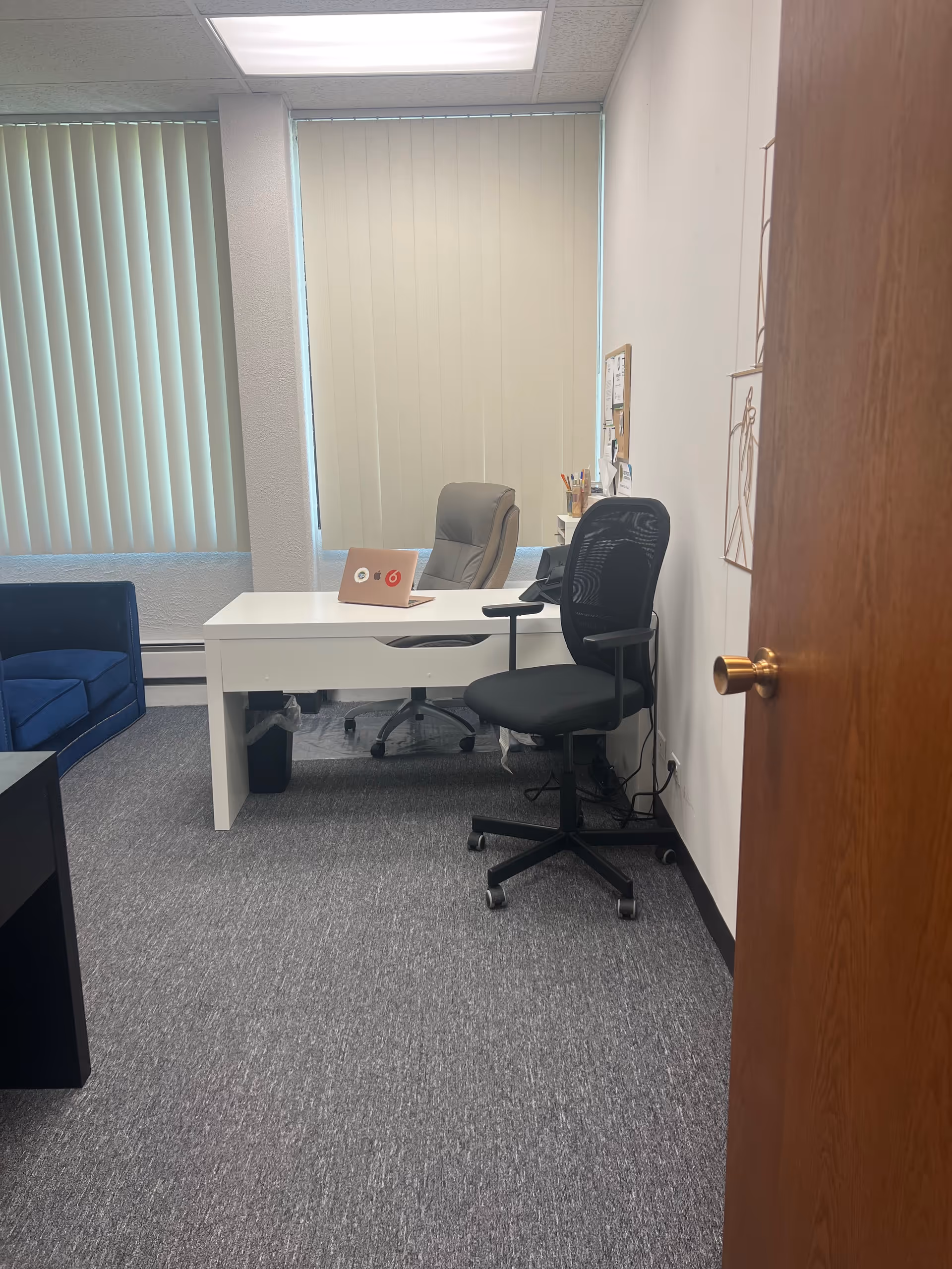 Office workspace with white desk, two chairs, vertical blinds, and an open door.