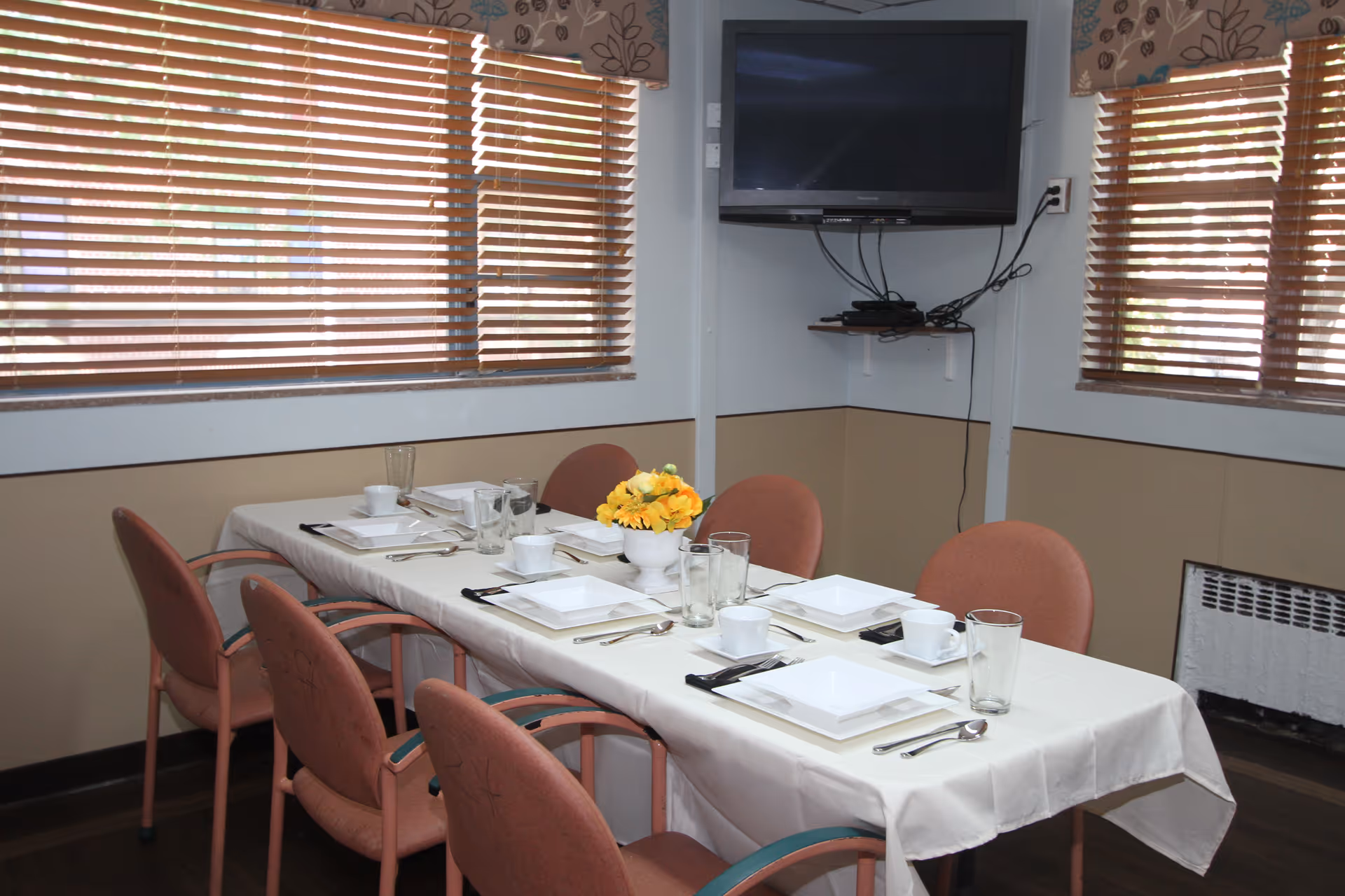 A dining room with a rectangular table covered with a white tablecloth, set with white square plates, cups, glasses, and silverware. Six pink cushioned chairs surround the table. A small vase with yellow flowers is placed in the center. The room has two windows with wooden blinds and a wall-mounted flat-screen TV above a small shelf with electronic devices.