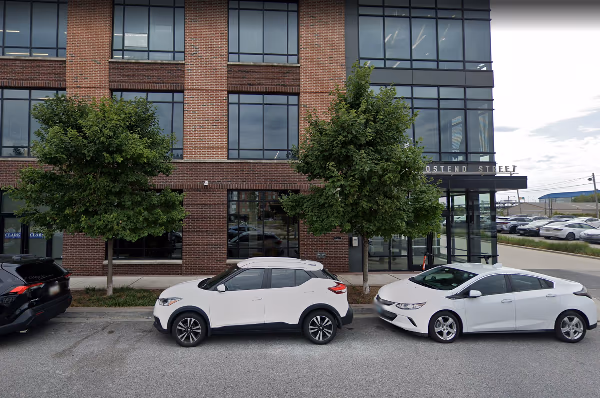 Brick building front with large windows, two trees, and parked cars along the street.