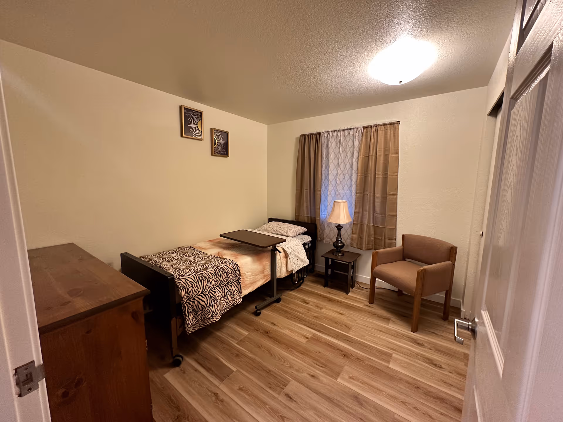 Cozy single bedroom with bed, dresser, nightstand lamp, window, and armchair.