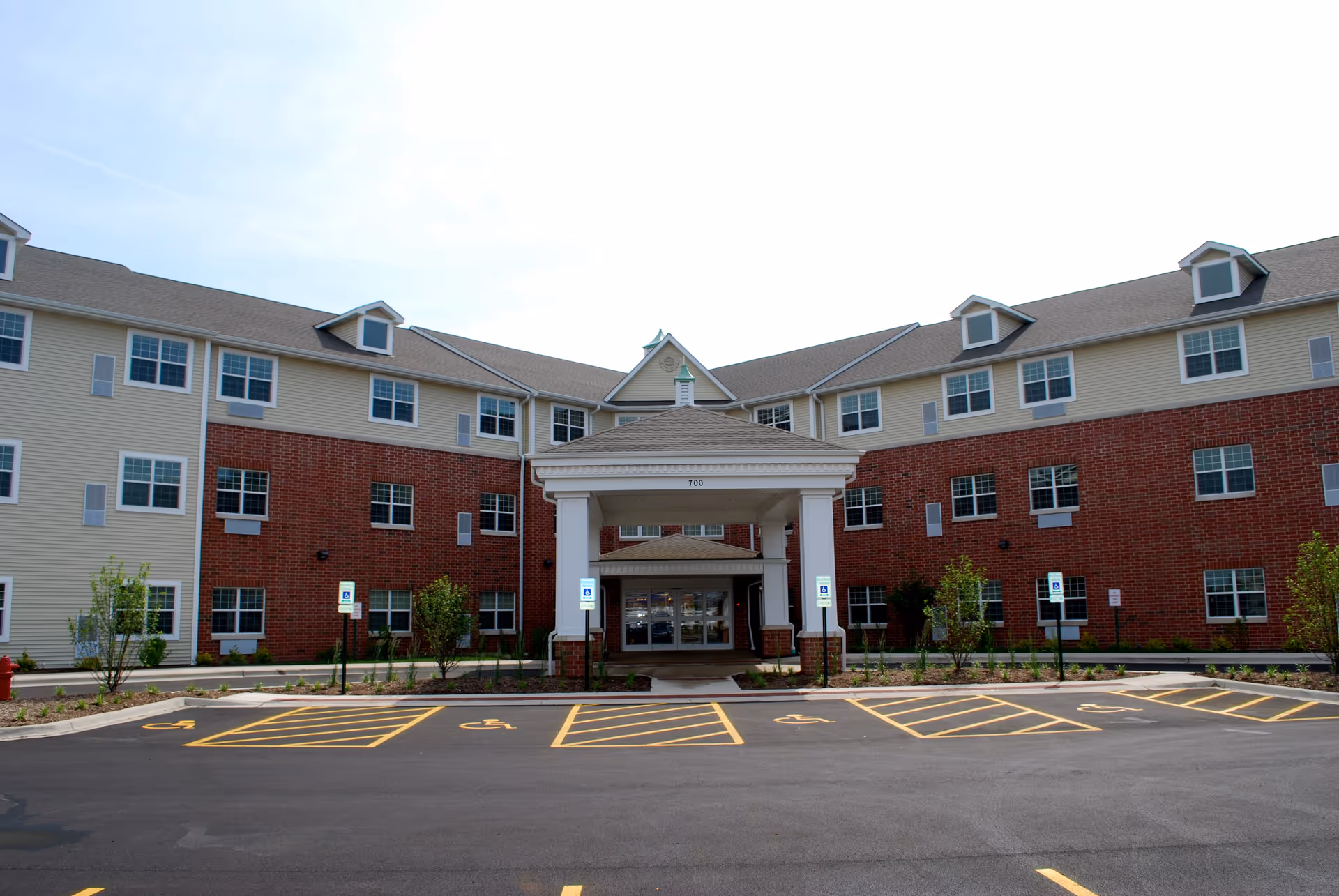 Front exterior view of a three-story senior living facility building with a covered entrance, multiple windows, and several handicap parking spaces in front.