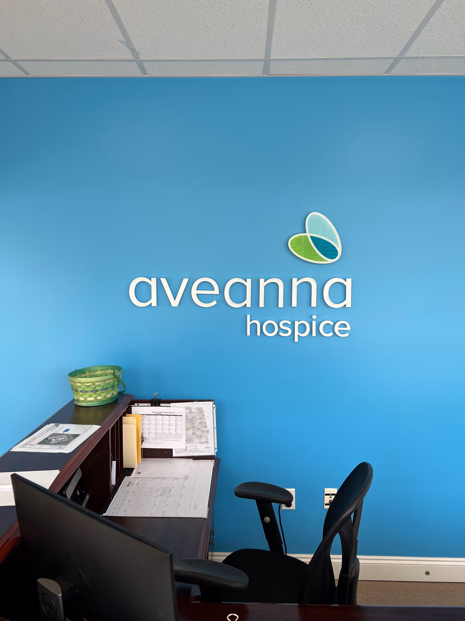 Blue wall with Aveanna Hospice logo, desk, papers, and office chair in reception area.