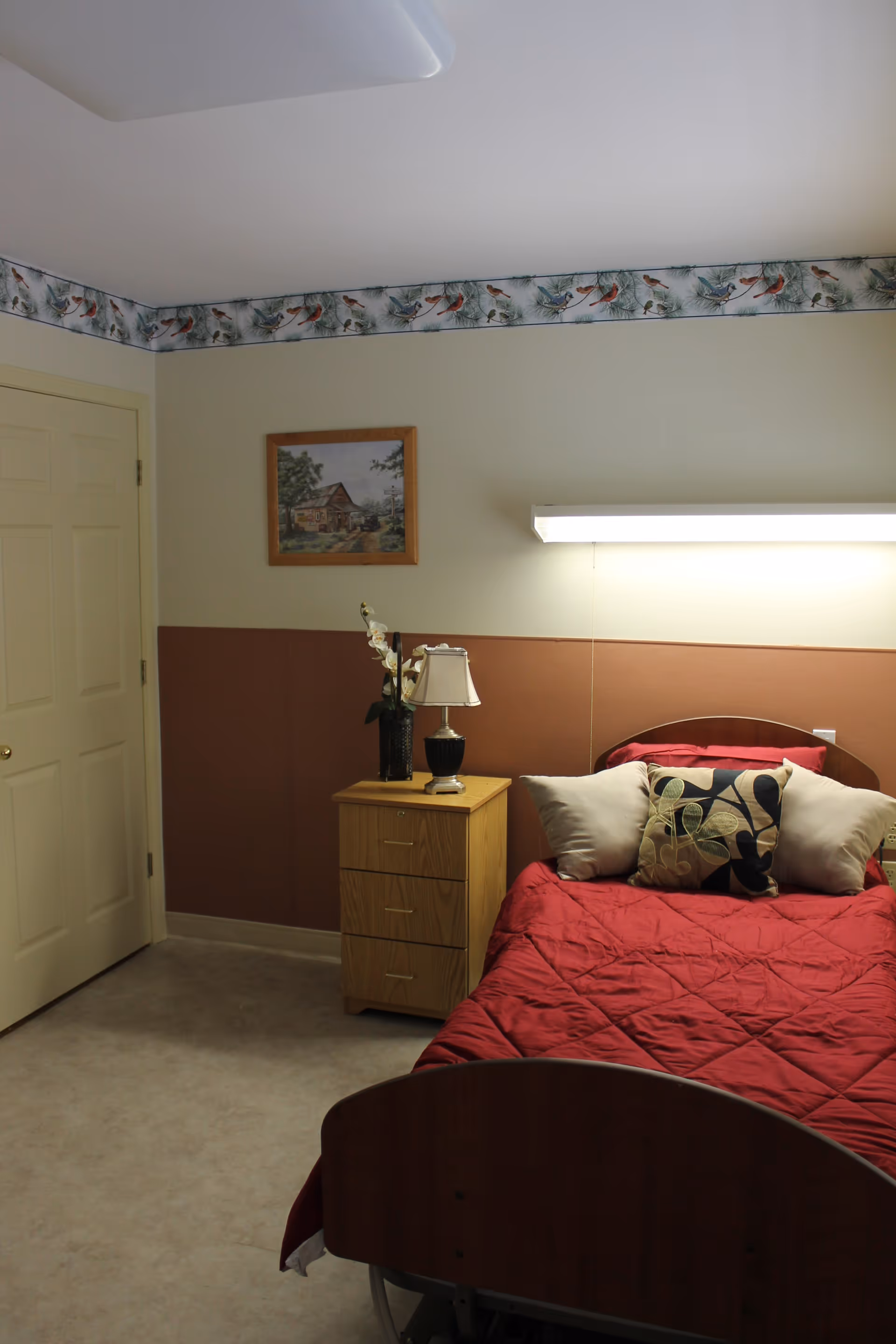 A bedroom in a healthcare facility with a bed covered in a red quilt and three decorative pillows. Next to the bed is a wooden nightstand with a lamp and a vase with flowers. A framed picture hangs on the wall above the nightstand. The walls are painted in two tones with a bird-themed wallpaper border near the ceiling.