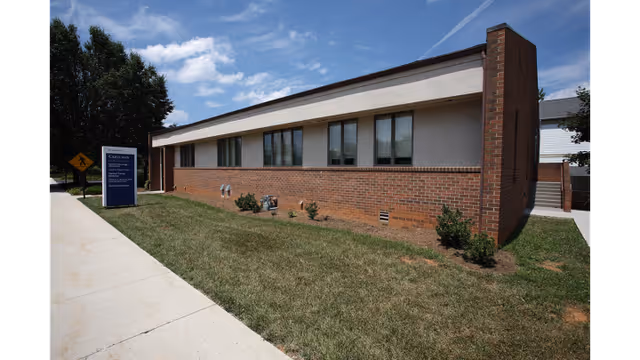 Single-story brick building with large windows, sidewalk, shrubs, and a blue information sign.