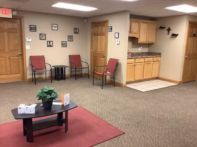 Interior lounge area with red chairs, small table, wall plaques, and kitchenette.