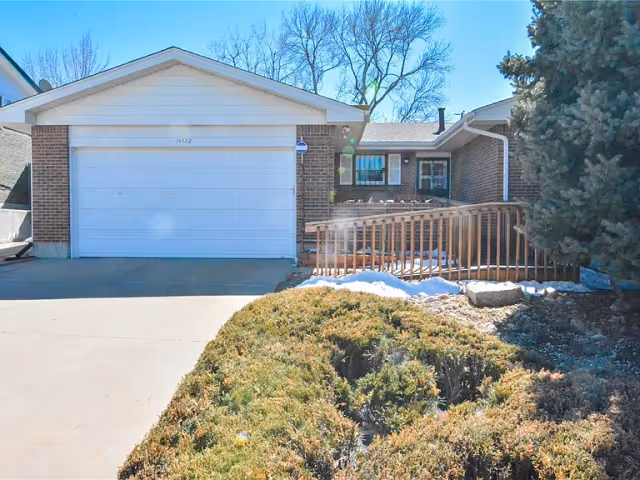 Single-story brick house front with a closed two-car garage, driveway, wooden accessibility ramp, and shrubs in the foreground.