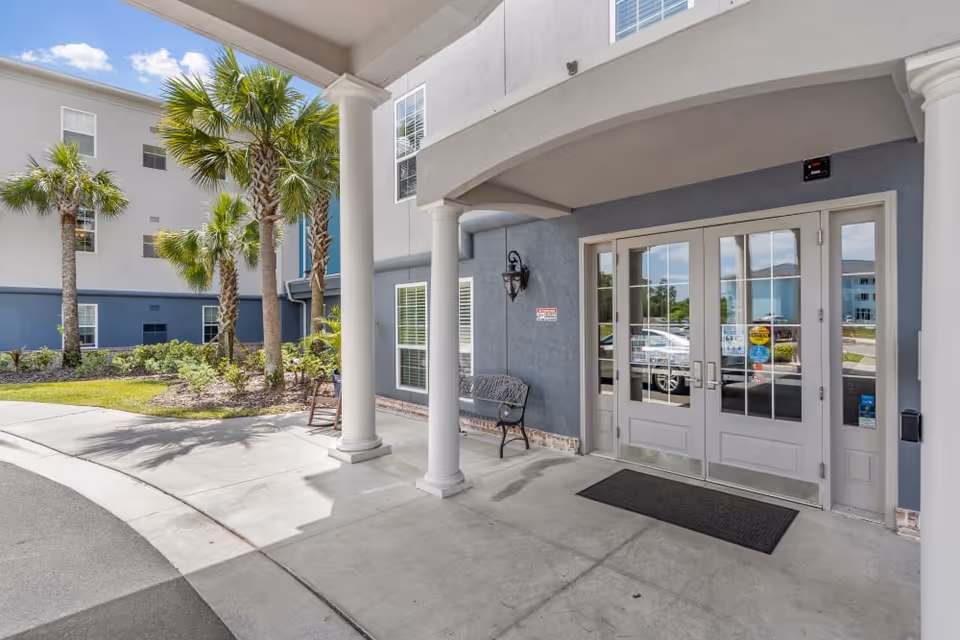 Entrance area of a senior living facility with double glass doors, two white columns, a black bench, and palm trees in the landscaped garden nearby under a blue sky with clouds.