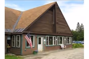 A brown wooden nursing home exterior with a steep roof, flag, and outdoor seating.