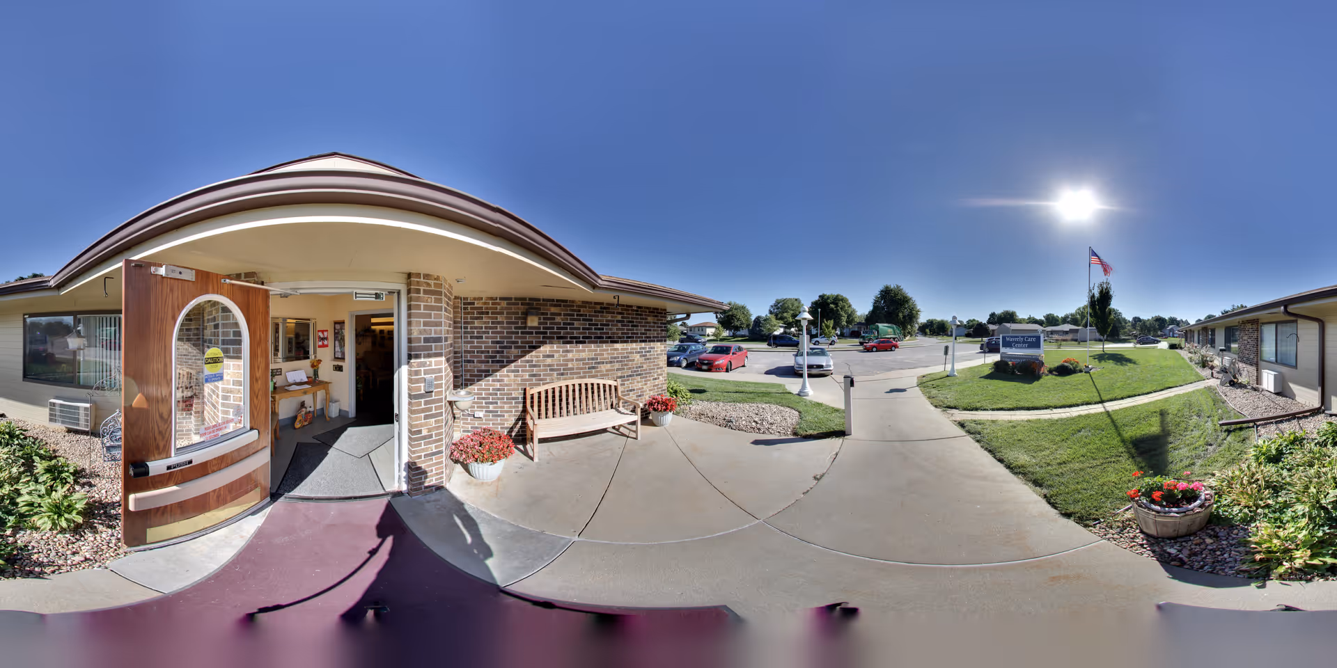 Wide-angle view of a senior living facility's front entrance with open double doors, a bench, planters, a driveway, flag, and parking area under a clear sky.