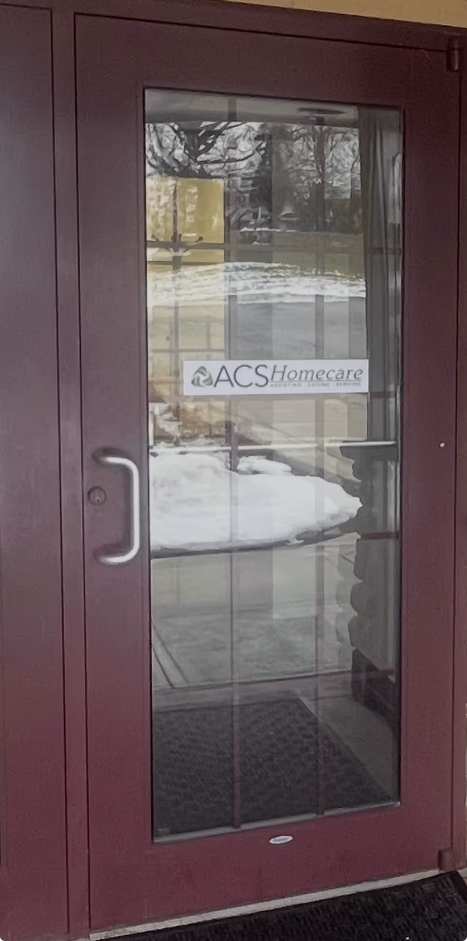 Maroon glass-paneled door entrance with ACS Homecare sign and snowy exterior reflections.